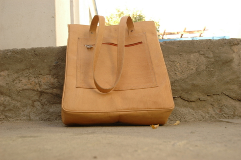 Tan leather tote bag with carved face detail on front, displayed against woven textile backdrop