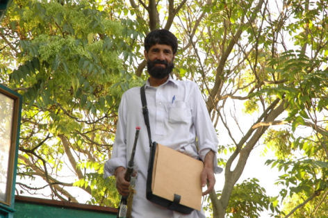 Man wearing light blue shirt and grey cardigan holding tan leather messenger bag under tree with green foliage