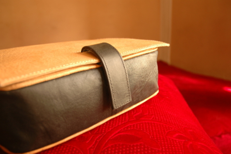 Tan leather bag with dark strap, displayed on wooden platform against warm orange wall and red surface