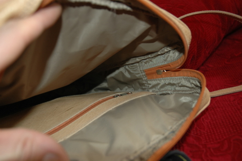 Hand holding open tan leather bag showing grey fabric lining with rust-colored seam detail against red background