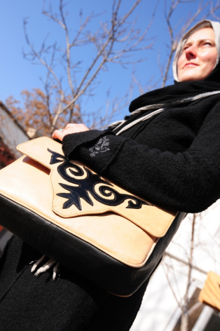 Frau in schwarzer Jacke hält eine cremefarbene Ledertasche mit schwarzem Ornament-Design, Außenaufnahme bei Tageslicht