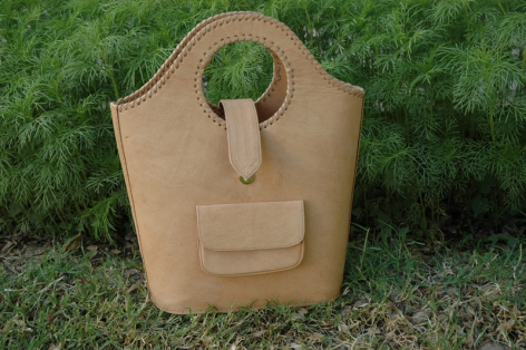 Beige Ledertasche mit Griff oben und aufgesetzter Vordertasche, frontal vor grünem Hintergrund