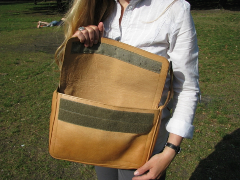 Hellbraune Ledertasche mit grünem Querstreifen, von vorne gezeigt, in den Händen einer Person vor grünem Hintergrund