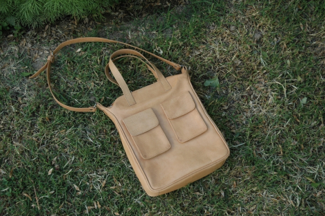 Beige Ledertasche mit quadratischen Fächern auf grünem Gras, Frontalansicht mit Schultergurt