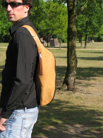 Person trägt handgefertigte cognacfarbene Ledertasche auf dem Rücken, Seitenansicht im Freien vor grünem Laub