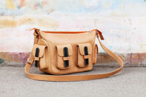 Tan leather shoulder bag with woven panel detail and brown leather handles, front view against brick wall