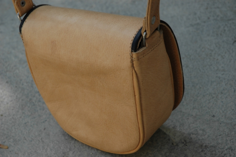 Sac à bandoulière en cuir beige clair, vue de trois quarts, avec rabat arrondi et détail de fermeture métallique
