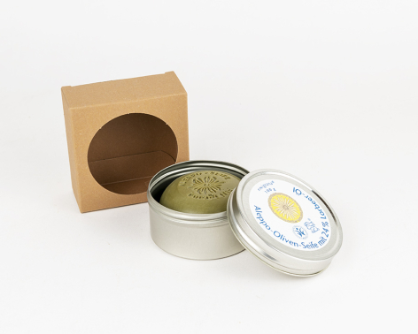 Metallic tin candle with yellow wax and clear lid, positioned beside kraft paper box with circular window