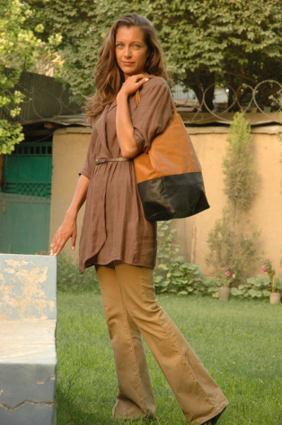 Woman in tan leather jacket and trousers with black crossbody bag standing against stone wall with greenery