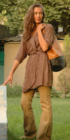 Woman wearing tan suede dress with fringe detailing and brown leather boots, standing outdoors with green background