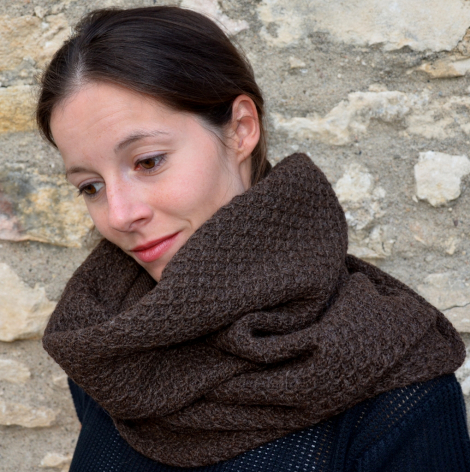 Woman wearing a chunky knit cowl neck scarf in charcoal brown, styled loosely over dark clothing against a stone wall