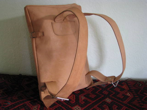 Tan leather backpack with rolled top closure and shoulder straps, displayed on dark fabric against white background