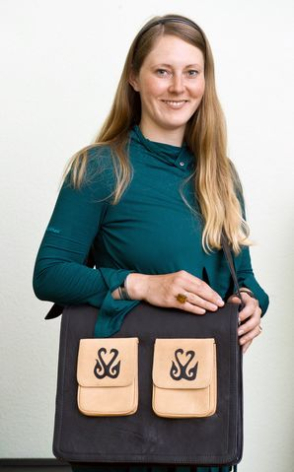 Woman with long brown hair in teal long-sleeved top holding black crossbody bag with cream geometric pockets
