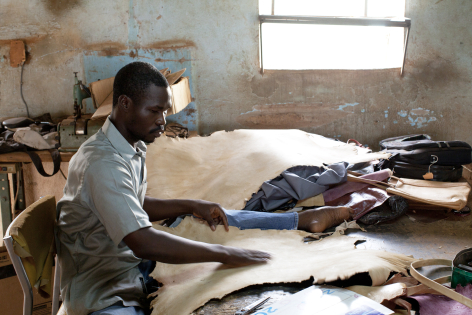 Artisan working at wooden table, hand-stitching dark blue leather in bright workshop with natural light from window