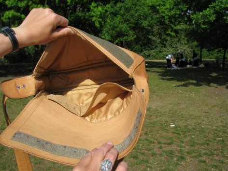 Brown leather tote bag held open showing tan canvas lining and interior pocket detail, outdoor setting with green grass