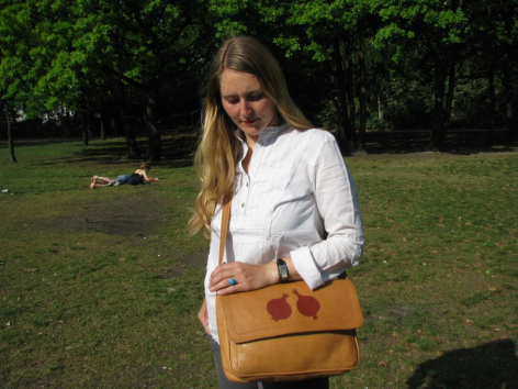 Woman wearing tan leather shoulder bag with front pocket, displayed on outdoor grass with trees in background