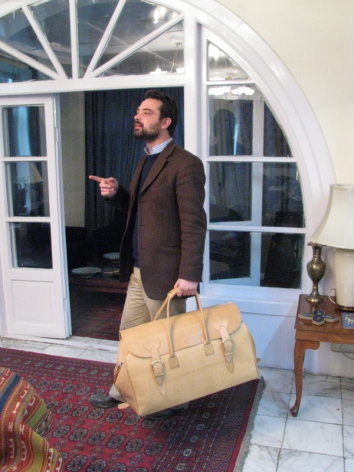 Man in brown jacket holding large tan leather duffel bag in front of arched doorway with white trim