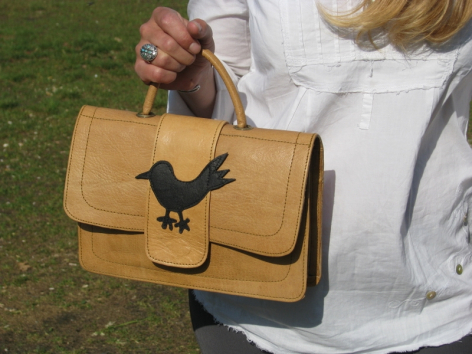 Tan leather shoulder bag with black bird silhouette emblem, held open to show interior compartment, front view