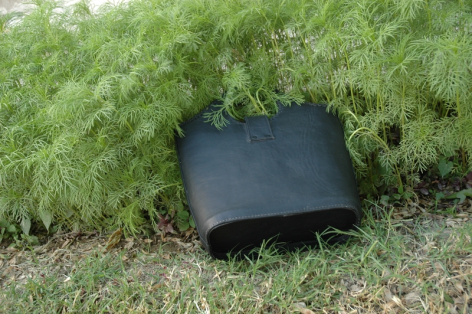 Black leather shoulder bag on green grass and foliage, top view showing closed flap