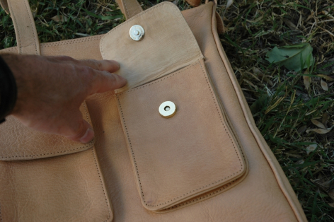 Brown leather bag with front pocket and metal snap closure, displayed on natural fabric background