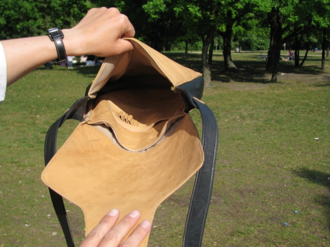 Hands holding open a tan leather bag outdoors, revealing the interior compartment in natural daylight