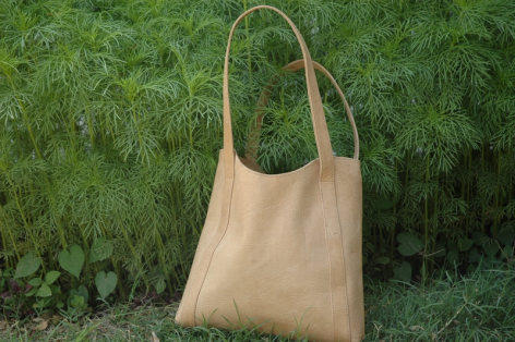 Cream leather tote bag with curved handles, displayed against green foliage background, front view