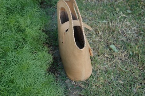 Tan leather bag with rounded shape and oval opening, displayed on grass from above