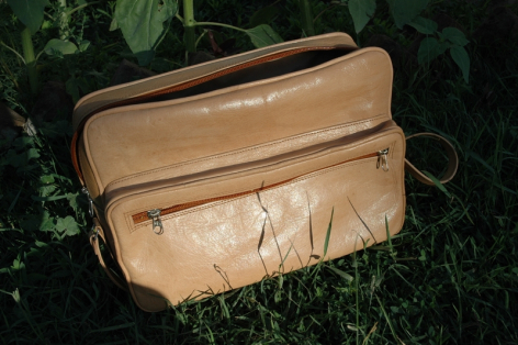 Tan leather messenger bag with front flap pocket and shoulder strap, displayed on grass