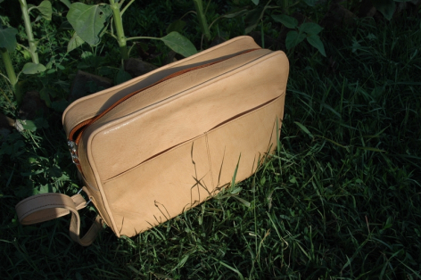 Tan leather satchel bag with dark top handle, displayed on grass in three-quarter view
