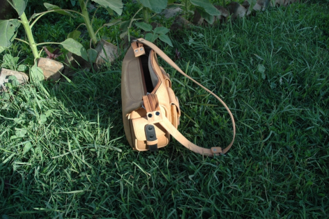 Tan leather crossbody bag with curved flap design, displayed flat on green grass, overhead view