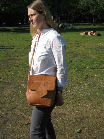 Woman wearing tan leather shoulder bag with front flap pocket, worn across body in outdoor park setting