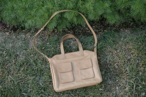 Beige leather tote bag with dual handles and front pocket detail, displayed on grass from above