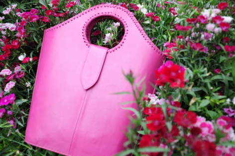 Pink fabric tote bag with circular leather handles displayed among blooming magenta and pink flowers in garden setting