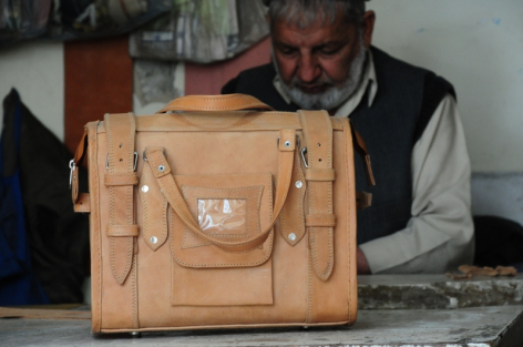 Craftsperson in vest examining tan leather bag with multiple pockets and straps during quality inspection