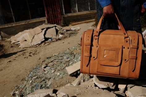 Tan leather travel bag with multiple compartments and handles on sandy ground, artisan workspace background