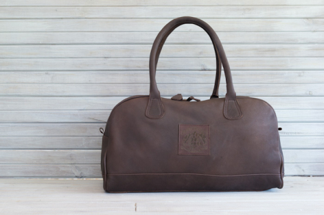 Brown leather duffel bag with dual handles, front view, displayed against white shiplap background