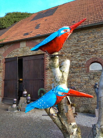 Two colorful macaw parrots perched on a wooden stand outside a stone building with terracotta roof tiles