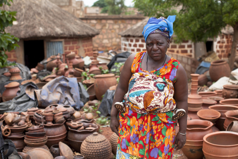 Woman in colorful patterned dress standing in brick production area with clay pots and earthen structures in background