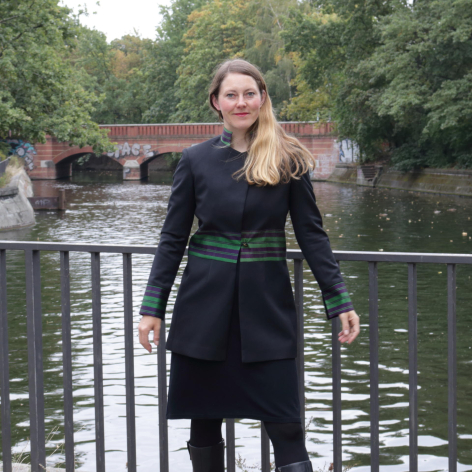 Woman in black coat standing on metal bridge over river with green trees in background