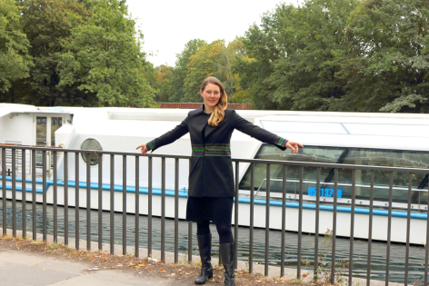 Woman in black outfit standing with arms outstretched on bridge overlooking water and green trees