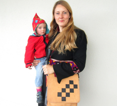 Woman holding young child in red jacket, wearing black top and holding tan leather bag with black geometric pattern