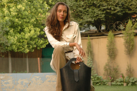 Frau in beiger Strickjacke und dunkler Hose hält eine braune Ledertasche mit Henkel vor sich