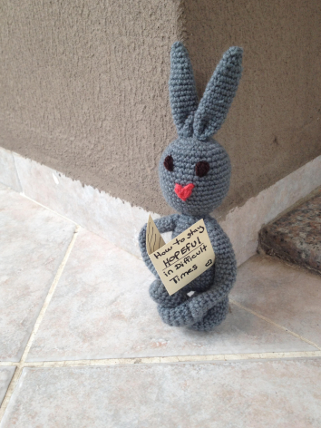 Grey knitted rabbit toy with long ears and red nose detail, sitting upright on tiled floor against beige wall