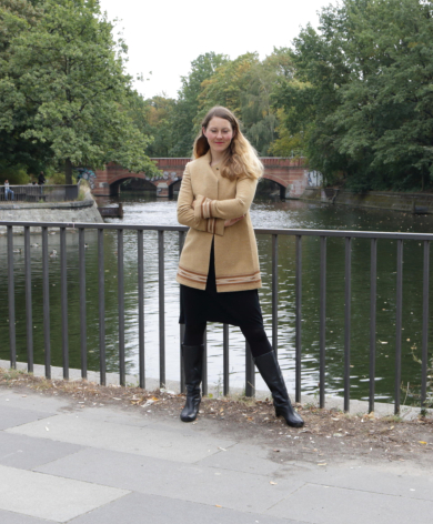 Woman wearing tan leather jacket standing on bridge railing with green trees and water in background