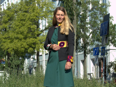 Woman in green dress holding small patterned brown leather crossbody bag against outdoor tree background