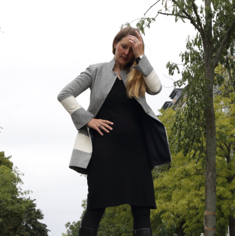Woman modeling black sleeveless dress with grey cardigan, standing outdoors among green foliage with hand on hip