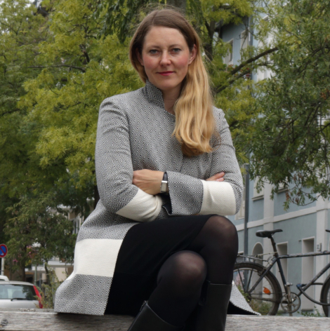 Frau sitzt auf einer Bank, trägt graue Strickjacke und schwarze Leggings, grüne Bäume im Hintergrund