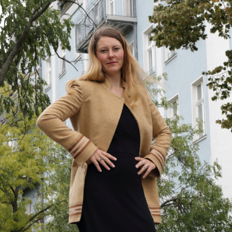 Woman wearing tan cardigan over black dress standing outdoors with green foliage and building in background
