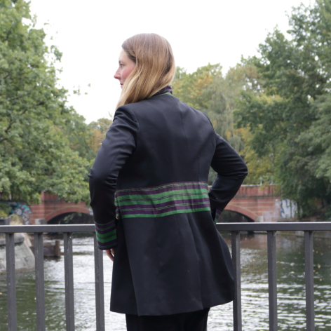 Frau von hinten in dunkelblauer Wolljacke mit grünem Tartanmuster um die Taille, an einem Geländer stehend