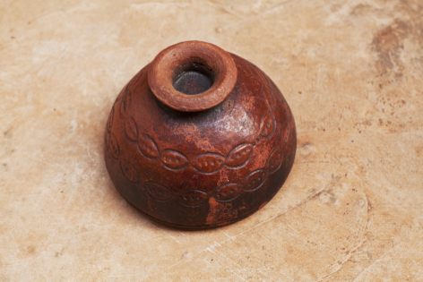 Rounded terracotta vessel with deep reddish-brown glaze and central opening, viewed from above on neutral surface